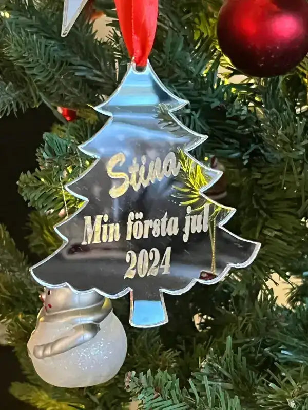 En "Julgranspynt min första jul" från Expresspresenter.se, graverad med "Stina Min första jul 2024", hänger i granen bredvid en snögubbe och röda kulor.