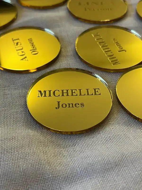 Flera runda, guldfärgade Placeringskort runda från Express-presenter visas på en ljus tygyta. På märket i fokus står det MICHELLE Jones med feta och regelbundna svarta bokstäver, och andra märken är elegant placerade i närheten.