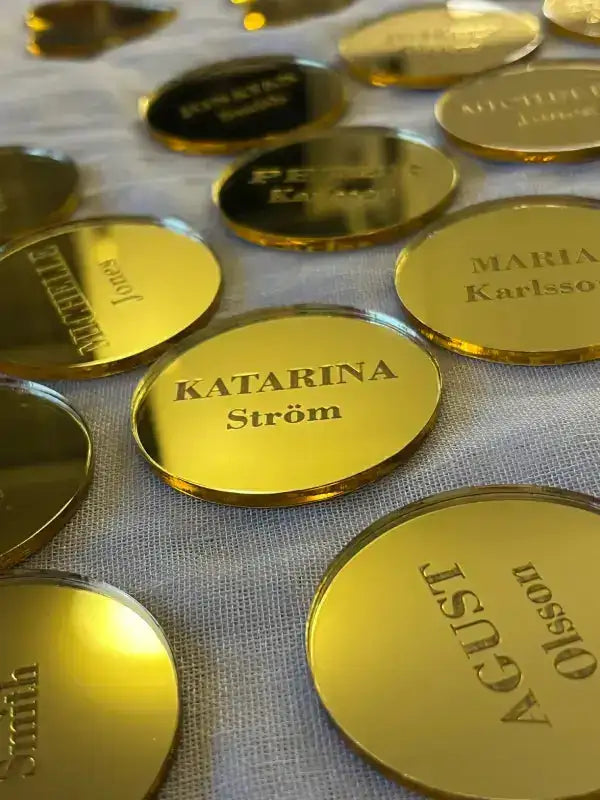 Flera runda, guldglänsande Placeringskort runda by Express-presenter med ingraverade namn som KATARINA Ström är utspridda på ett vitt tyg. Dessa personliga bordsplaceringar fångar ljuset och vissa namn är delvis synliga.