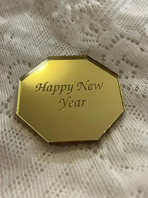 Express-presenter Bordsdekoration Happy New Year är en guldglänsande, åttkantig spegel akrylplatta med texten "Happy New Year", som är placerad på ett vitt mönstrat tyg för en dekorativ touch.