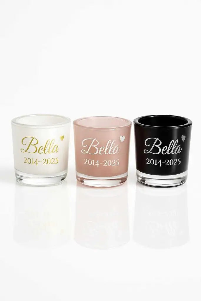 Tre ljuslyktor ur Express-presenters kollektion "Minneslykta namn & Årtal", märkta Bella 2014-2025 med guld- och vitskriven text på rosa, svart och vitt glas, placerade sida vid sida. Perfekt för värmeljus.