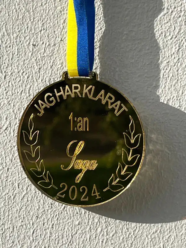 På en vit vägg hänger en "Medalj | 1:an" från Express-presenter med ett blått och gult band. Denna spegelplast guld medalj med namn har inskriptionen "JAG HAR KLARAT 1:an Saga 2024" med lagerbladsmönster på varje sida.