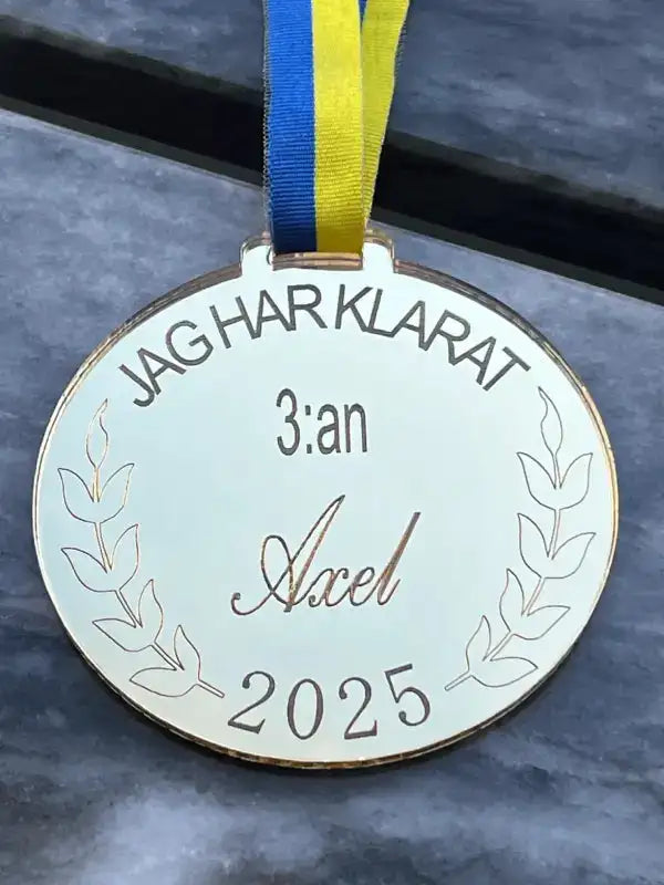 Medalj | 3:an från Express-presenter är en guldfärgad spegelblank medalj med "JAG HAR KLARAT 3:an Axel 2025" och lagergravyr, på ett blågult band, som visas vilande på en gråmarmorerad yta.