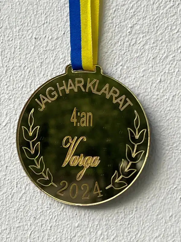 Medalj | 4:an by Express-presenter i spegelblank guldplast med blågult band, med gravering "JAG HAR KLARAT 4:an Varga 2024" och lagergrenar, placerad mot vit vägg.