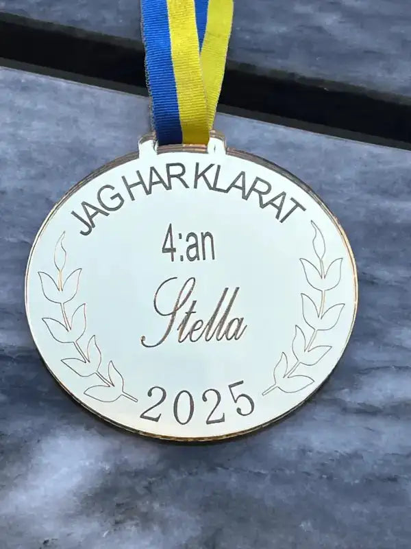 Medalj | 4:an från Express-presenter har blågult band, lagerkvistar, spegelplast guldfinish, personlig gravyrtext "JAG HAR KLARAT 4:an Stella 2025" och visas på en grå marmorliknande yta.