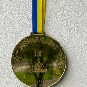 Medalj | 8:an från Express-presenter i spegelplast guld med blå/gul rosett hänger mot en vit vägg. Medaljen är graverad med "JAG HAR KLARAT 8:an Albin 2021" och dekorerad med lagerkvistar.