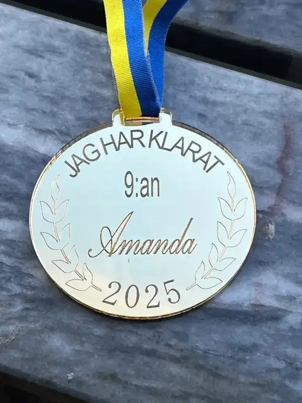 Expressens presentatörsmedalj 9:an i spegelplast guld, med blågult band och egenhändigt utförd svensk gravyr "JAG HAR KLARAT 9:an Amanda 2025" ovanför lagergrenar, vilar på en gråmarmorerad yta.