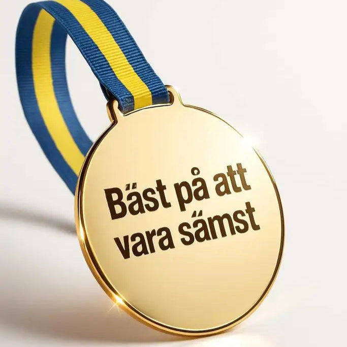 Medalj | Bäst på att vara sämst" från Express-presenter är en guldmedalj i spegelplast med blågult band, graverad med den svenska frasen "Bäst på att vara sämst".