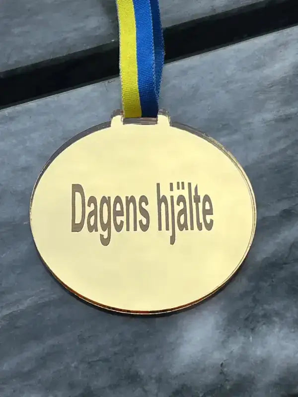 Medalj | Dagens hjälte by Express-presenter består av en guldmedalj med ett blågult sverigeband, graverad med "Dagens hjälte" på svenska, som visas på en grå yta.