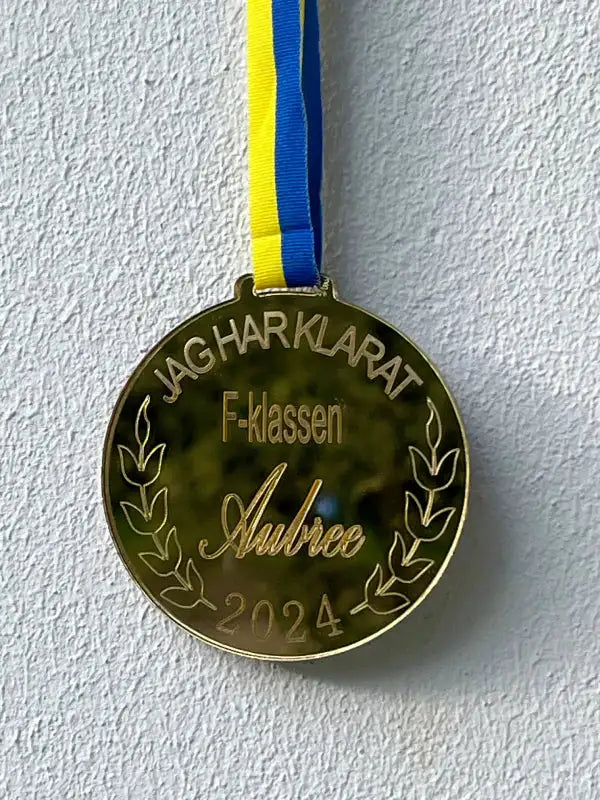 En personlig Medalj | F-klassen från Express-presenter med blått och gult band, graverad med "JAG HAR KLARAT F-klassen Aubree 2024" och lagerkvistar, hänger mot en ljusstrukturerad vägg.