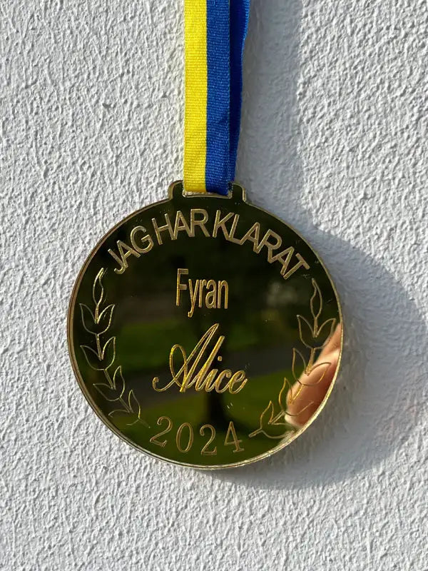 En Medalj | Fyran från Express-presenter hänger från ett blågult band mot en vit vägg. Medaljen har personlig gravyr: "JAG HAR KLARAT Fyran Alice 2024" och dekorativa lagerkvistar.