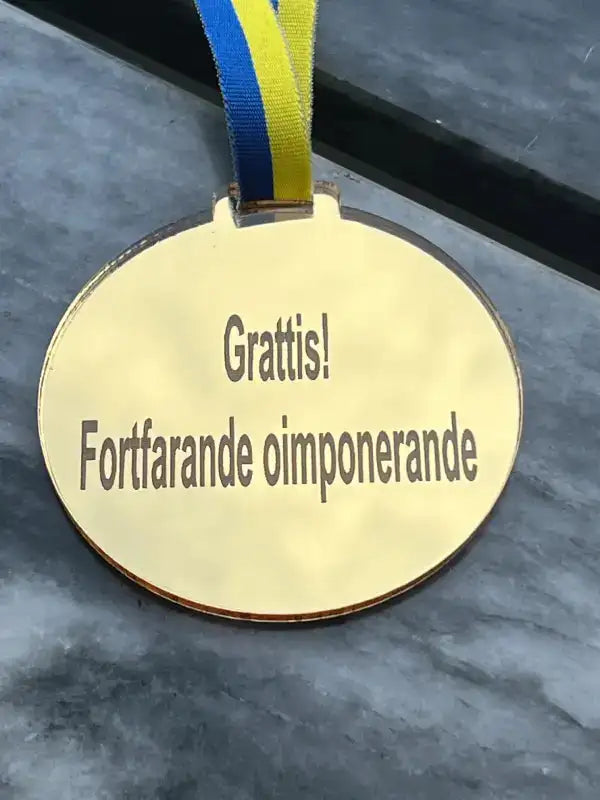 En Medalj | Grattis från Express-presenter vilar på en grå yta med ett blågult sverigeband. Den 3mm tjocka medaljen är graverad med "Grattis! Fortfarande oimponerande.