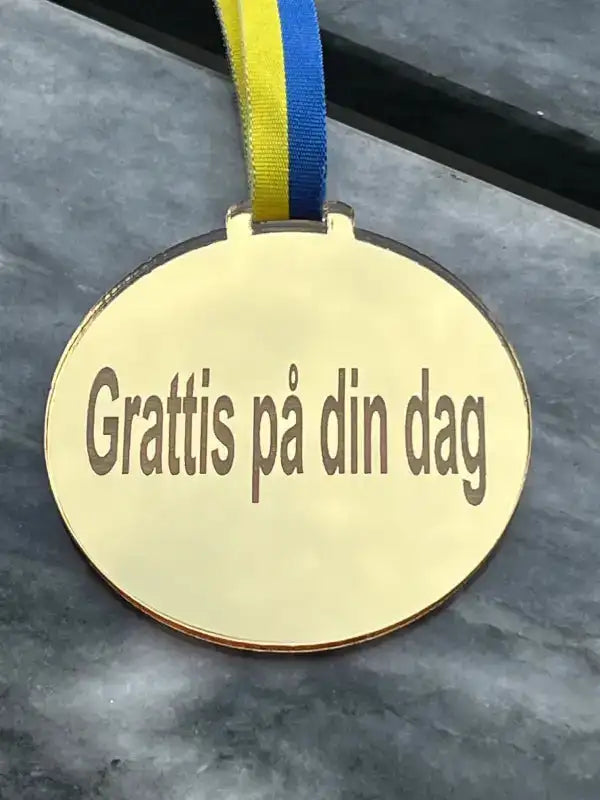 Medaljen Grattis på din dag by Express-presenter är en guldfärgad spegelplastmedalj med blågult sverigeband och graveringen "Grattis på din dag", på en grå marmoryta.