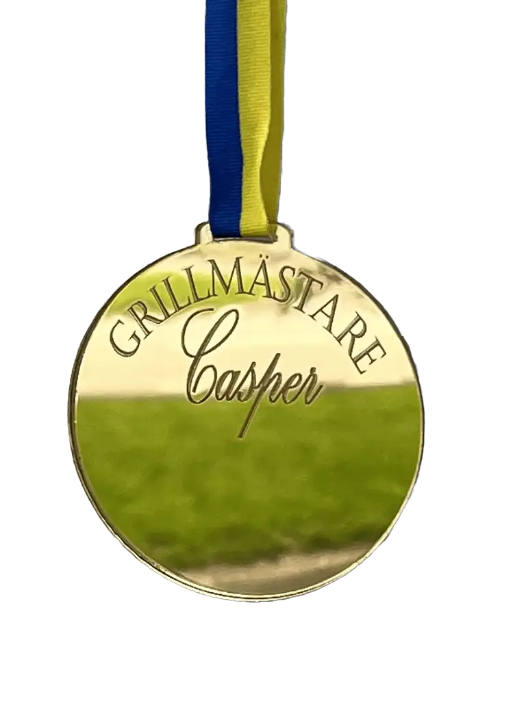 Medaljen Grillmästare från Express-presenter, graverad med "GRILLMÄSTARE Casper", hänger i ett blågult band och skimrar när den fångar ljuset över ett grönt, gräsbevuxet fält.
