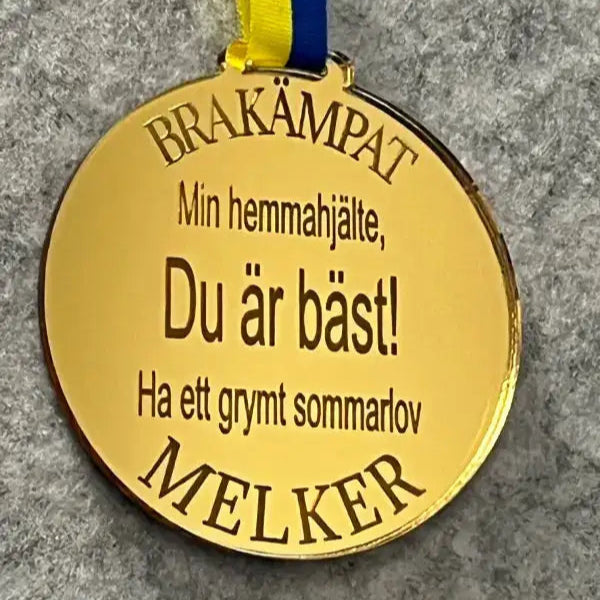 Expressens presentatörsmedalj | Min hemmahjälte Du är bäst! är en guldfärgad spegelplastmedalj med blått och gult Sverigeband, svensk text: "BRA KÄMPAT. Min hemmahjälte, du är bäst! Ha ett grymt sommarlov. MELKER", mot grå bakgrund.