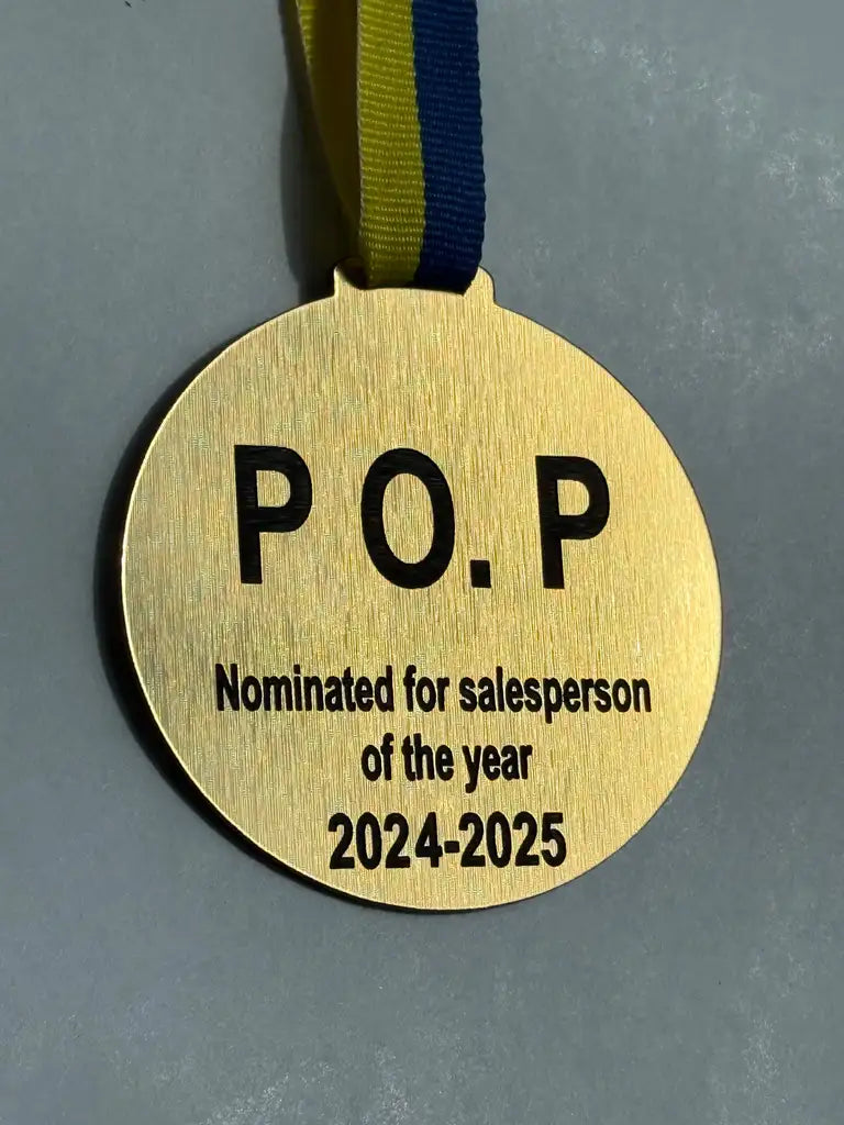 En guldmedalj, Nominerad för bästa by Express-presenter, med blågult band och graveringen "P.O.P Nominerad till årets säljare 2024-2025", är det perfekta kickoff-priset i graverad akryl.