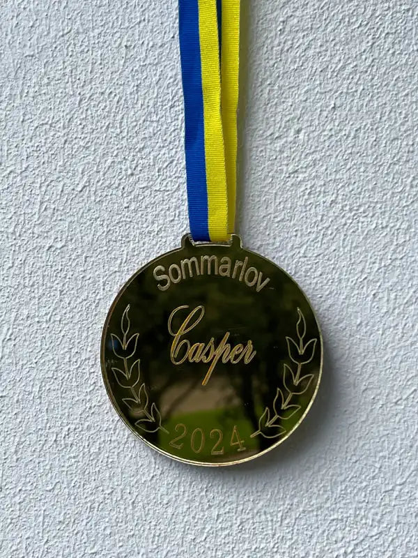 Mot en vit vägg hänger en Medalj | Sommarlov by Express-presenter, med spegelplast guld, ett blågult band och graverat med "Sommarlov", "Casper", "2024" och lagerkvistar.