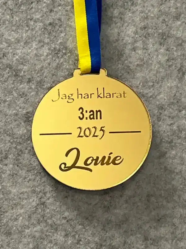 Express-presenter Medalj | jag har klarat 3:an - en graverad medalj med Sverigeband i blått och gult och texten "Jag har klarat 3:an 2025 Louie" på svenska, visas på en grå yta.