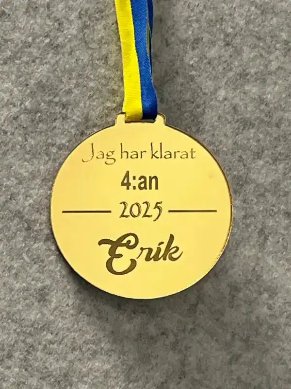 Express-presenter Medalj | jag har klarat 4:an: Guldmedalj i spegelplast med blågult Sverigeband. Graverad "Jag har klarat 4:an 2025 Erik." Grå filtbakgrund. Bra årskurs 4 minnesmärke.