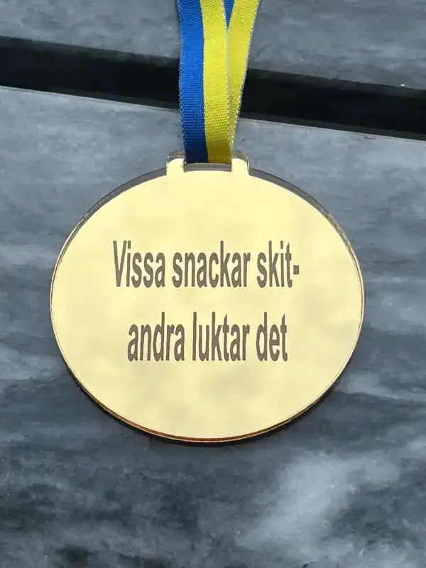 En Medalj | rolig by Express-presenter med ett blågult sverigebandsband vilar på en grå yta, graverad med "Vissa snackar skit-andra luktar det.