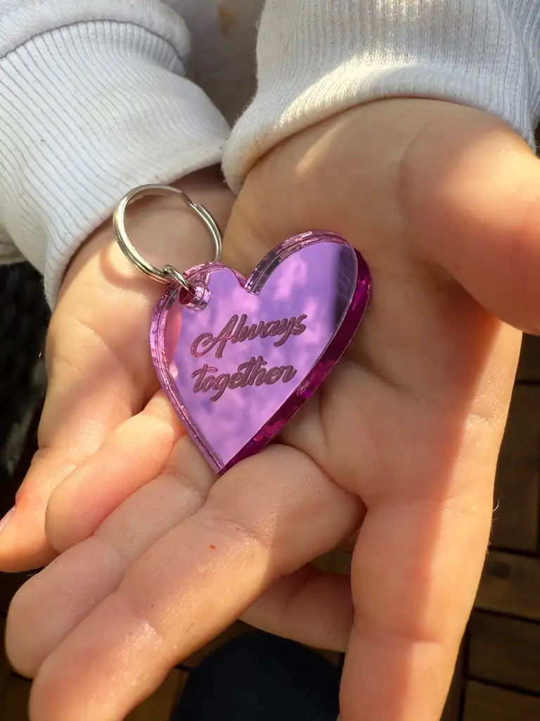 En person i vit långärmad skjorta håller i Express-presenter Nyckelring - Always together (Rosa), en 4x3cm hjärtformad nyckelring i rosa spegelplast (3mm) med "Always together" ingraverat i kursiv stil.