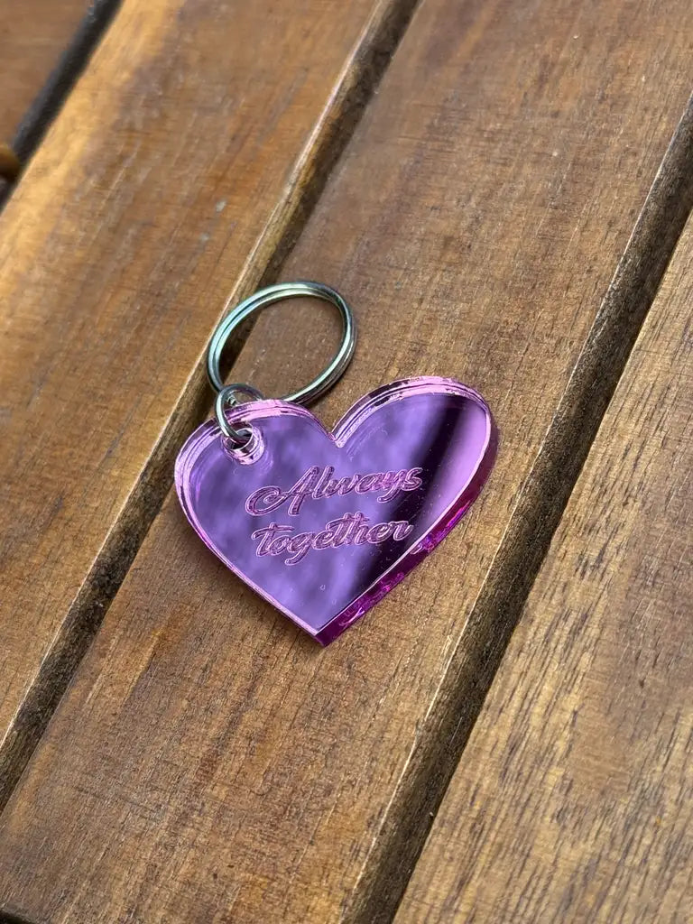 Nyckelringen - Alltid tillsammans (Rosa) från Express-presenter är en rosa, hjärtformad nyckelring i spegelplast (4x3 cm, 3 mm tjock) med graveringen "Alltid tillsammans", som vilar på en träyta.