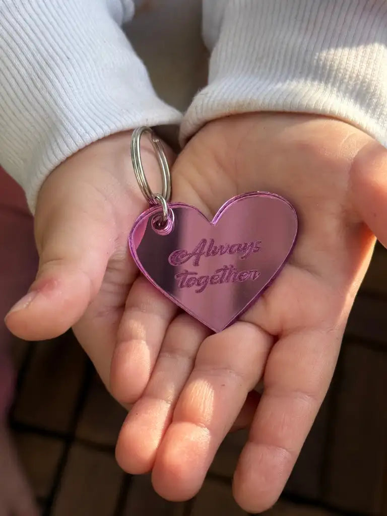 Ett barn i vit långärmad skjorta håller i Expressens Nyckelring - Alltid tillsammans (Rosa), en 4x3 cm, 3 mm tjock hjärtformad nyckelring i spegelplast med kursiv gravyr "Alltid tillsammans".