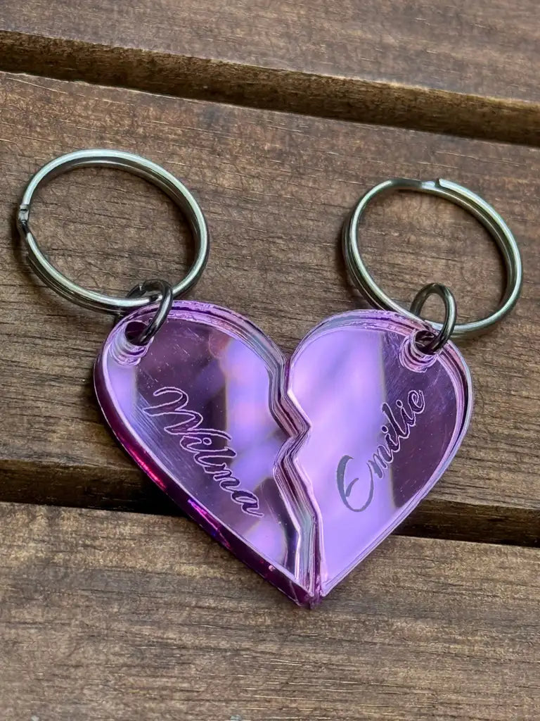 Två nyckelringar Nyckelring - Halvt hjärta i spegelplast (rosa) från Express-presenter, vardera 4x3cm och graverade med namn, bildar ett förenat rosa hjärta när de sätts ihop. De genomskinliga berlockerna visas på en träyta.
