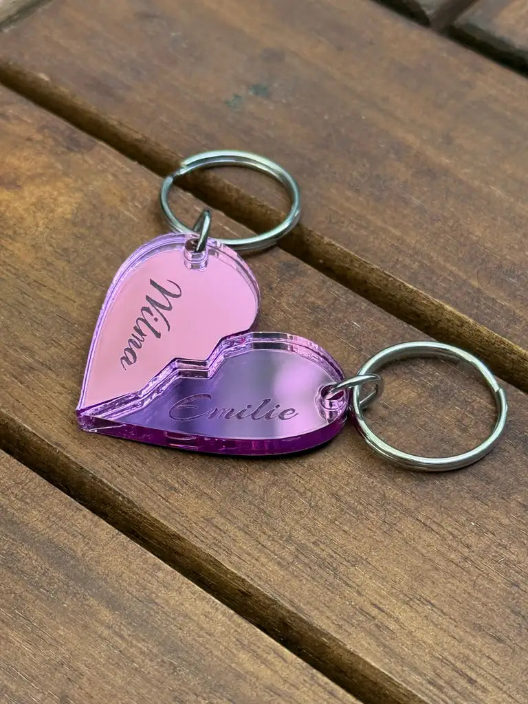 Två Nyckelring - Halvt hjärta i spegelplast (rosa) av märket Express-presenter, vardera 4x3 cm, vilar på trä. Nyckelringarna i rosa spegelplast har ingraverade namn och sätts ihop till ett hjärta.