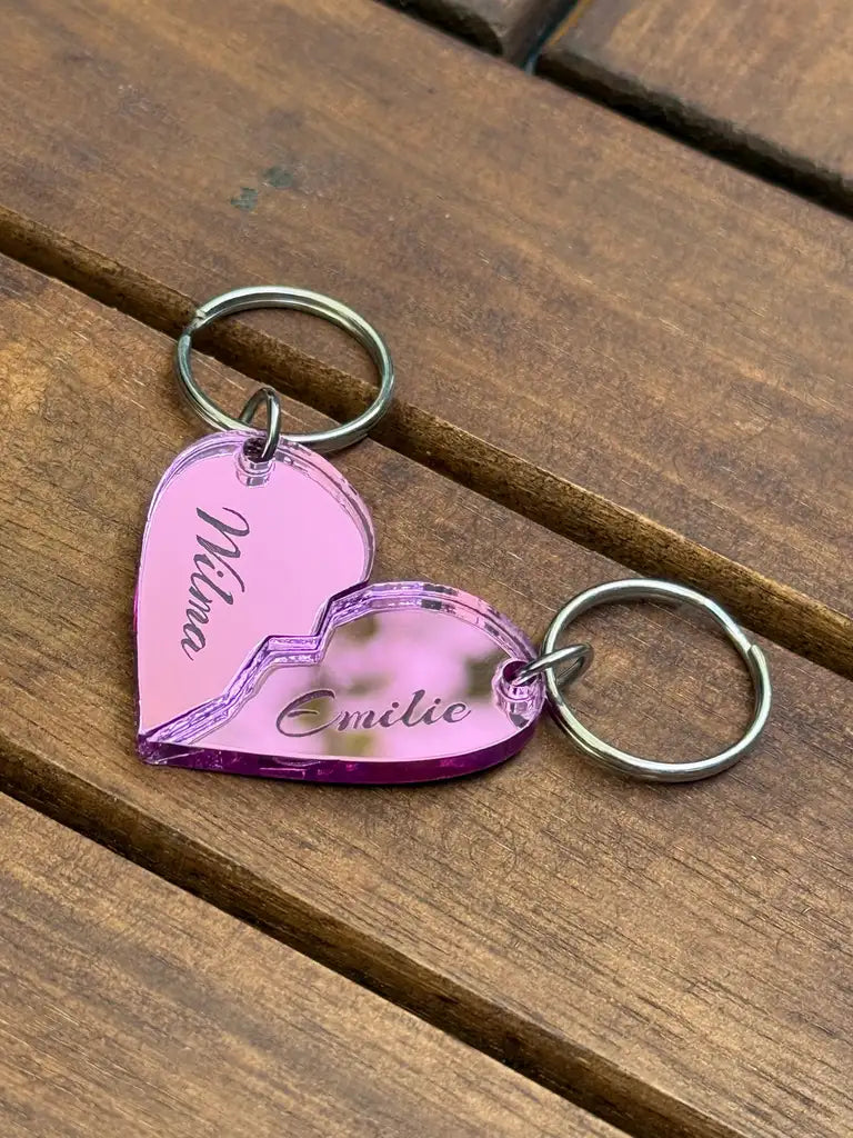 Två rosa "Nyckelring - Halvt hjärta i spegelplast (rosa)" av Express-presenter, vardera 4x3 cm, bildar ett hjärta med namnen Milina och Emilie. Båda halvorna har nyckelringar och visas på en träyta.