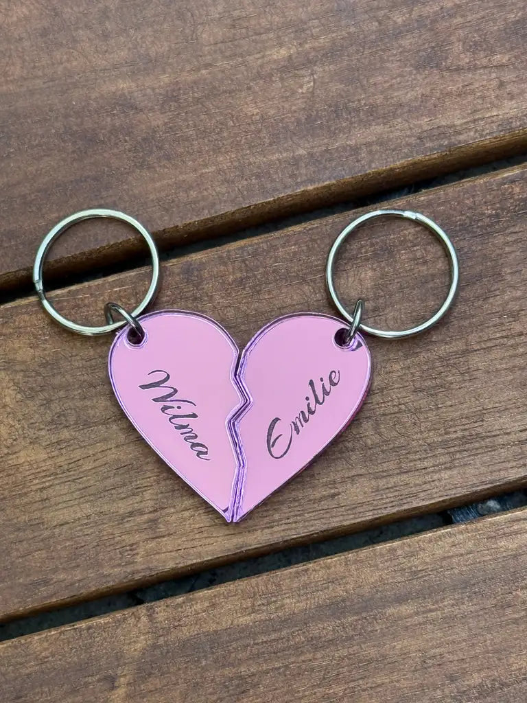 Två Nyckelring - Halvt hjärta i spegelplast (rosa) från Express-presenter, vardera 4x3cm med namnen Wilma och Emilie i hjärtform, visas separerade på en träyta.