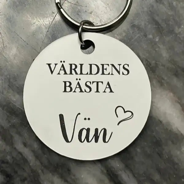 Nyckelringen Världens bästa vän (vit) från Express-presenter är en rund vit nyckelring med "världens bästa vän" och ett hjärta på, sett på en grå marmoryta - perfekt som present till din bästa vän.