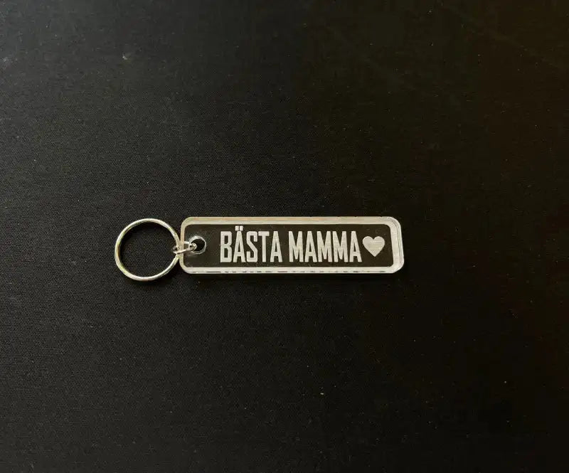 Nyckelring | Bästa mamma från Express-presenter: rektangulär akryl, svart botten med vit text "BÄSTA MAMMA" och hjärta. Placeras på mörk yta. Perfekt present - graverad baksida gör den extra personlig.