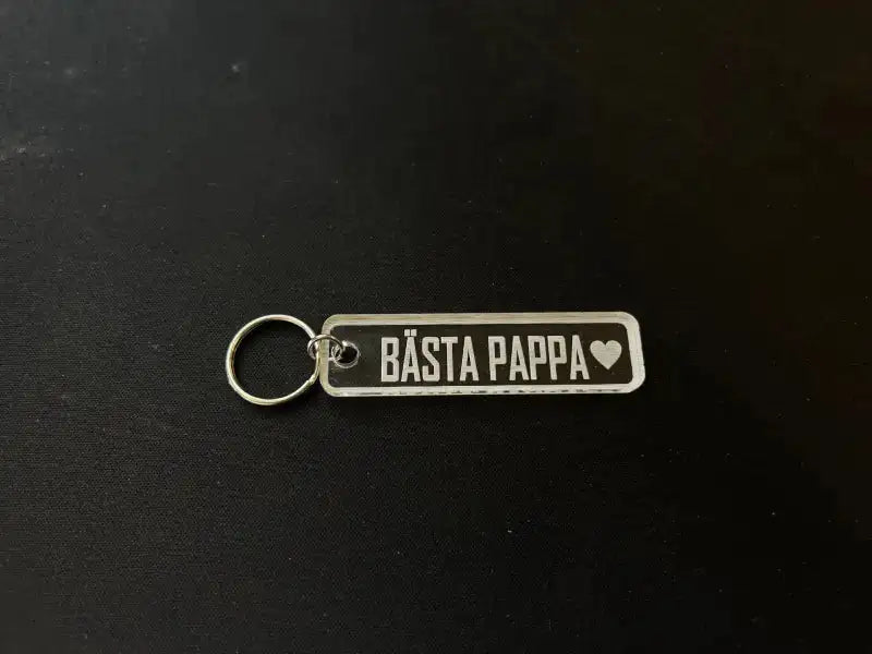 Nyckelring | Bästa pappa från Express-presenter - rektangulär nyckelring med metallring, graverad på fram- och baksidan, med texten BÄSTA PAPPA och hjärta på framsidan, visad på svart underlag.
