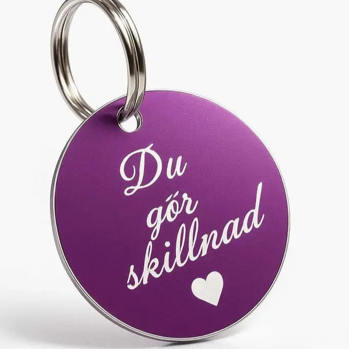 Den runda, mörklila Express-presenter-nyckelringen "Du gör skillnad" har silverkanter, metallring, är 40mm bred och dekorerad med svensk text samt ett vitt hjärta.