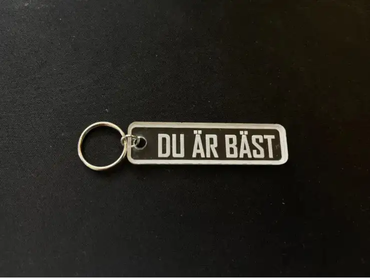 Express-presentatören Nyckelring | Du är bäst är en rektangulär nyckelring i akryl med metallring, graverad med den svenska frasen "DU ÄR BÄST", vilande på en svart yta.