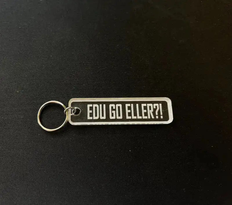 En rektangulär Nyckelring | Edu go eller by Express-presenter med metallring visas på en svart yta. Nyckelringen i 3 mm akryl har texten "EDU GO ELLER?!" i feta vita bokstäver på svart bakgrund.
