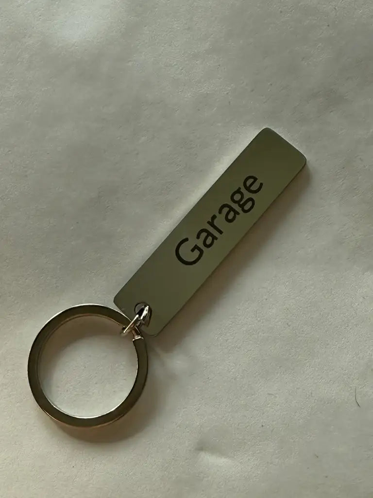 En Nyckelring | Garage från Express-presenter med en rektangulär metalltag lasermärkt med "Garage", ligger på en ljus yta.
