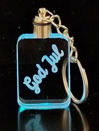 Nyckelringen God Jul från Expresspresenter.se är en rektangulär nyckelring i akryl med graveringen "God Jul" och blå LED-upplysta kanter, fäst på en nyckelring och kedja i metall, mot en svart bakgrund.