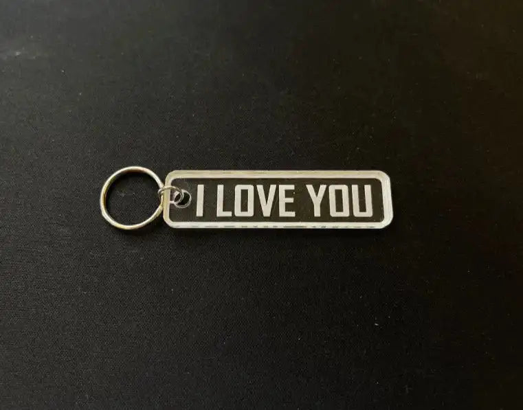 Express-presenter Nyckelring | I love you är en rektangulär nyckelring i akryl med texten "I LOVE YOU" i feta vita bokstäver på mörk bakgrund, placerad på en svart yta.