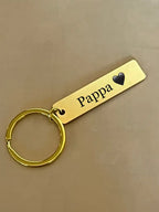 Nyckelringen Pappa från Expresspresenter.se är en nyckelring i guld med en rektangulär tagg i rostfritt stål, graverad med "Pappa" och ett svart hjärta, på en beige yta.