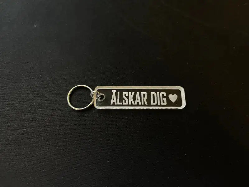 Nyckelringen Älskar dig by Express-presenter är en nyckelring i klar akryl med en metallring, graverad med "ÄLSKAR DIG" och en hjärtsymbol, på en svart yta.