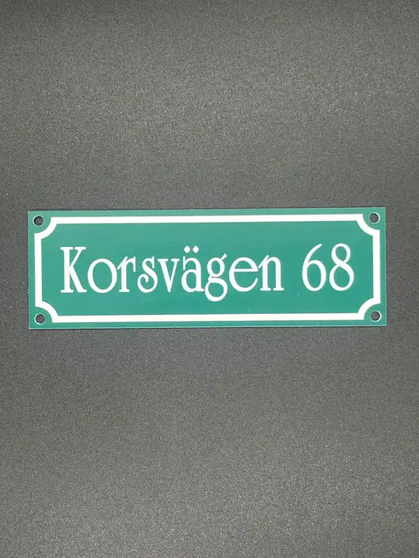 Express-presenter Postlådeskylt 150x50mm är en grön rektangulär skylt med vit text "Korsvägen 68", en vit kant och fyra hörnhål för montering.