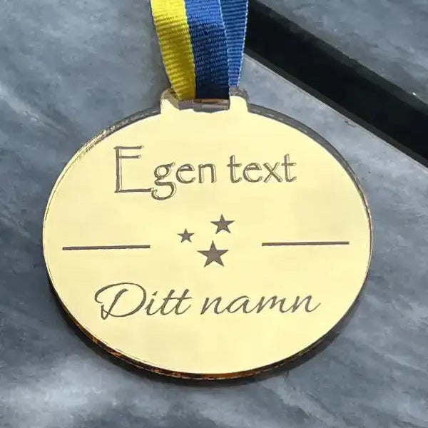Medalj | Egen text by Express-presenter är en guldmedalj med 3mm blågult sverigeband, graverad med "Egen text" och "Ditt namn" ovan och under tre stjärnor, utställd på en grå marmoryta.
