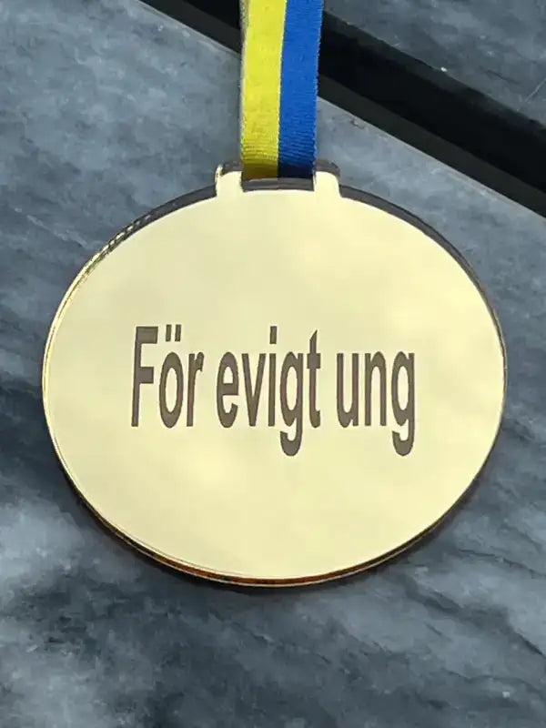 Medaljen För evigt ung by Express-presenter är en guldmedalj med ett blågult sverigeband och budskapet "För evigt ung" ingraverat på blank spegelplast, placerad på en grå marmoryta.