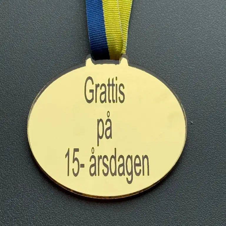 Express-presenter Medalj | Grattis på 15-årsdagen är en oval medalj (70 x 3 mm) i spegelplast guld, med ett blågult band och inskriptionen "Grattis på 15-årsdagen" mot en mörk bakgrund.