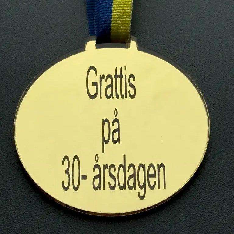 Medalj | Grattis på 30-årsdagen by Express-presenter är en guldoval spegelplast guldmedalj med blågult band, storlek 70 x 3 mm, graverad med "Grattis på 30-årsdagen", visad på en mörk yta.