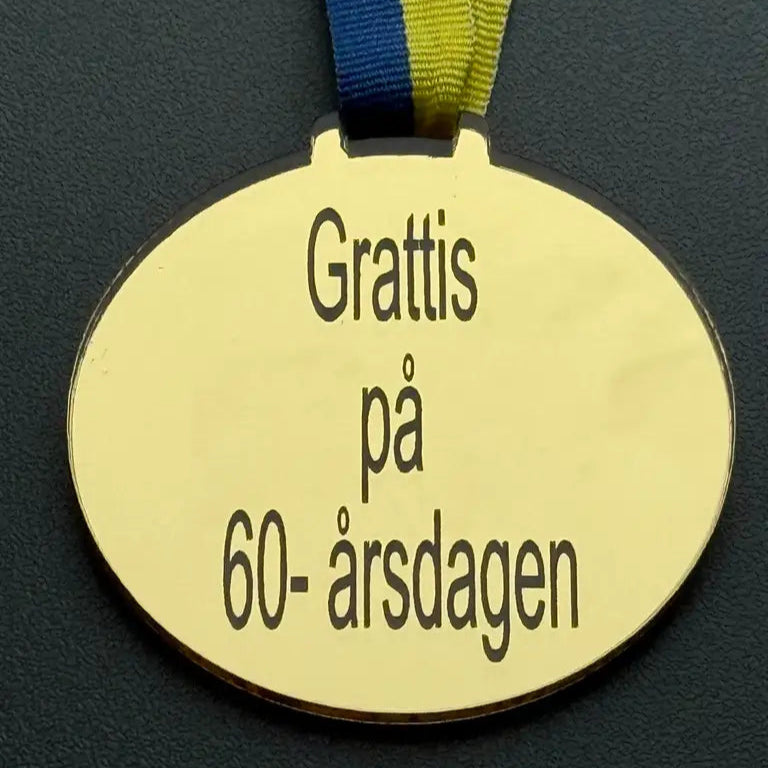 Express-presenterar Medalj | Grattis på 60-årsdagen: Guldfärgad oval medalj i spegelplast med blågult band och svensk text "Grattis på 60-årsdagen".