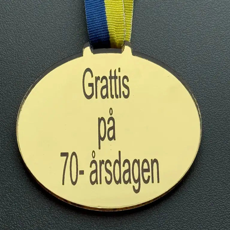 Medalj | Grattis på 70-årsdagen från Express-presenter, i spegelplast guld (70 x 3 mm) med blågult band och graverad svensk text, visas mot en mörk bakgrund.