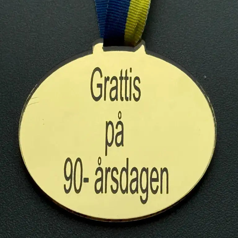 Express-presenters "Medalj | Grattis på 90-årsdagen" är en oval medalj (70 x 3 mm) i spegelplast guld, med blågult band och inskription, här avbildad på en mörk yta.
